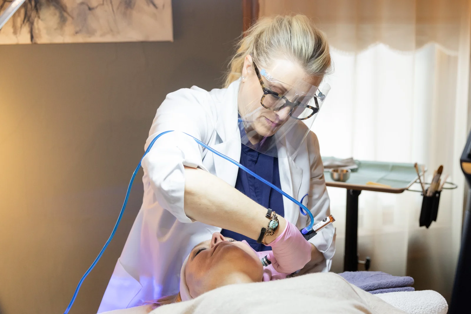 Clinician in a white lab coat, glasses and a face shield guiding a HydraFacial handpiece connected to blue tubing along a reclined client's jawline during a HydraFacial treatment.